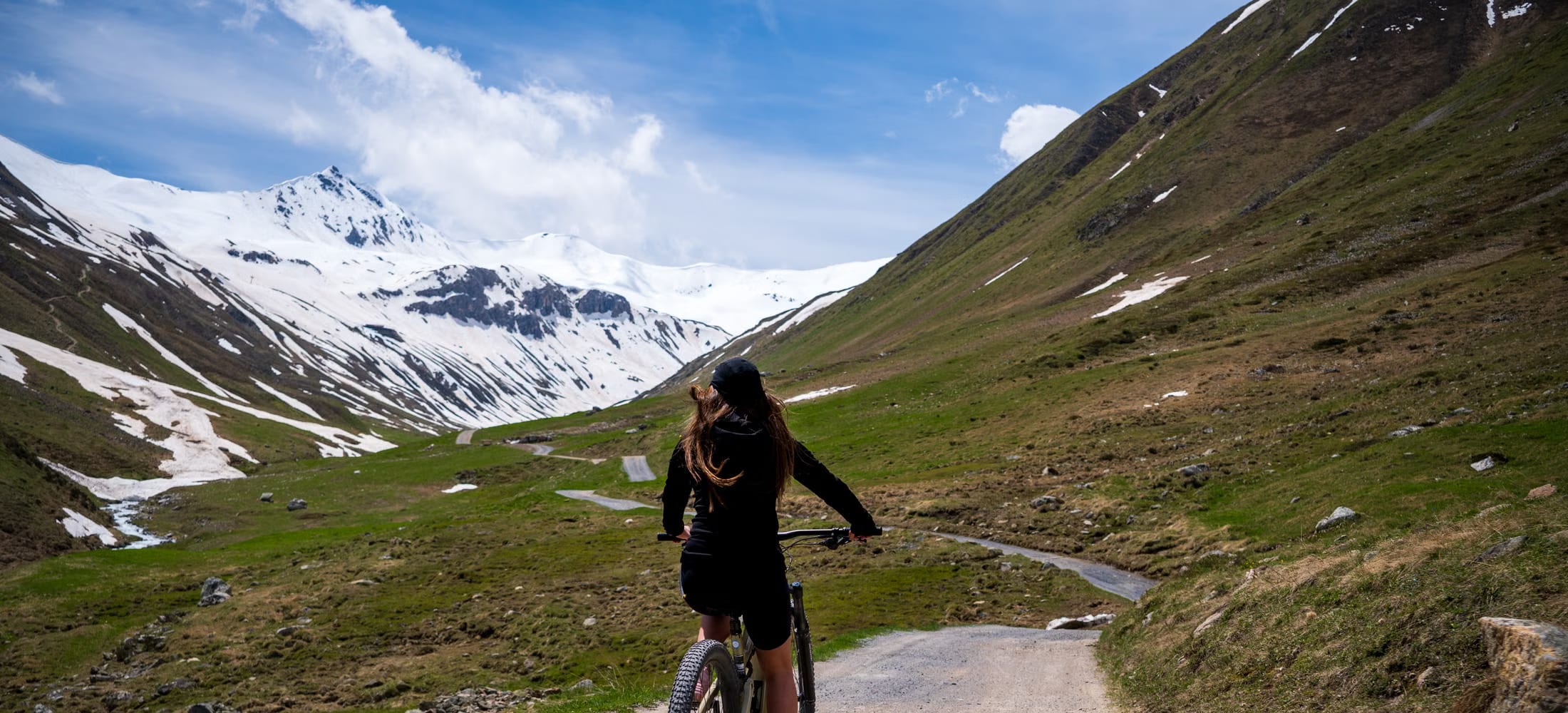 Radfahrerin auf einem Schotterweg durch ein grünes Hochtal, im Hintergrund ein schneebedeckter Bergkamm unter blauem Himmel