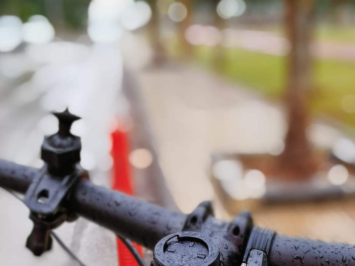 Nahaufnahme eines regennassen Fahrradlenkers mit leerer Gerätehalterung, dahinter ein unscharfer Radweg