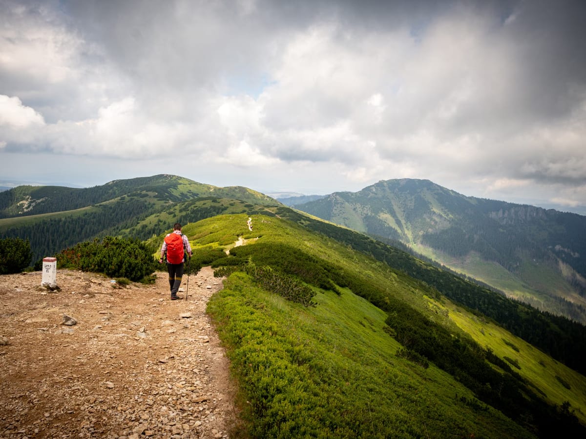 Wanderer mit rotem Rucksack auf einem schmalen Gratweg über bewaldeten Bergrücken unter dichter Wolkendecke